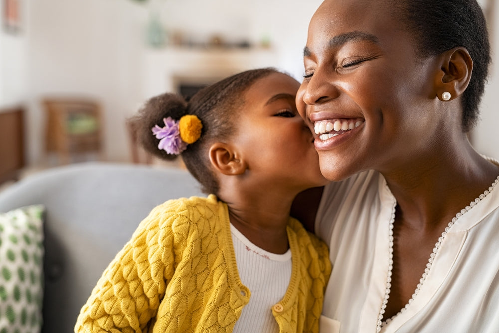 Mom smiling with young daughter who is giving her a kiss on the cheek.